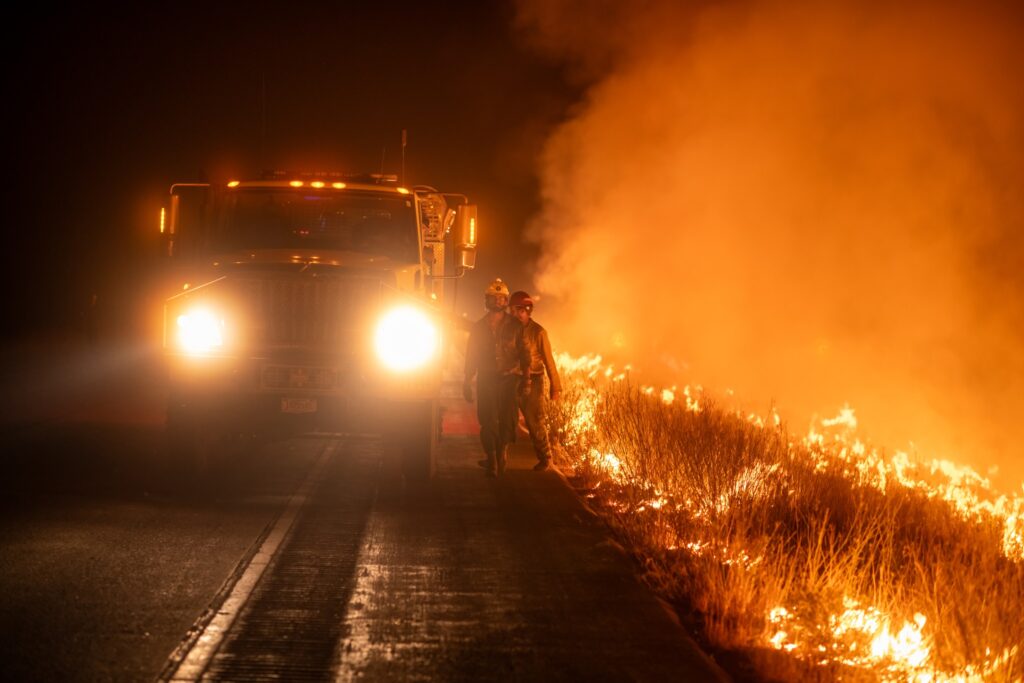 Two firefighters walk alongside a firetruck and next to a wildfire
